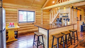 Interior de una cabaña de troncos con zona de cocina, suelo de madera, barra con taburetes y una escalera.