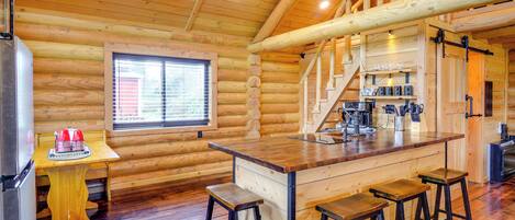 A log cabin interior with a kitchen area, wooden flooring, a bar with stools, and a staircase.
