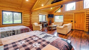 Interior de una cabaña de troncos con dos camas, una chimenea, un televisor y un ventanal grande.