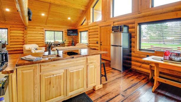Interior de una cabaña de troncos con una mesa de comedor de madera, una isla de cocina, un refrigerador y un televisor montado en la pared.