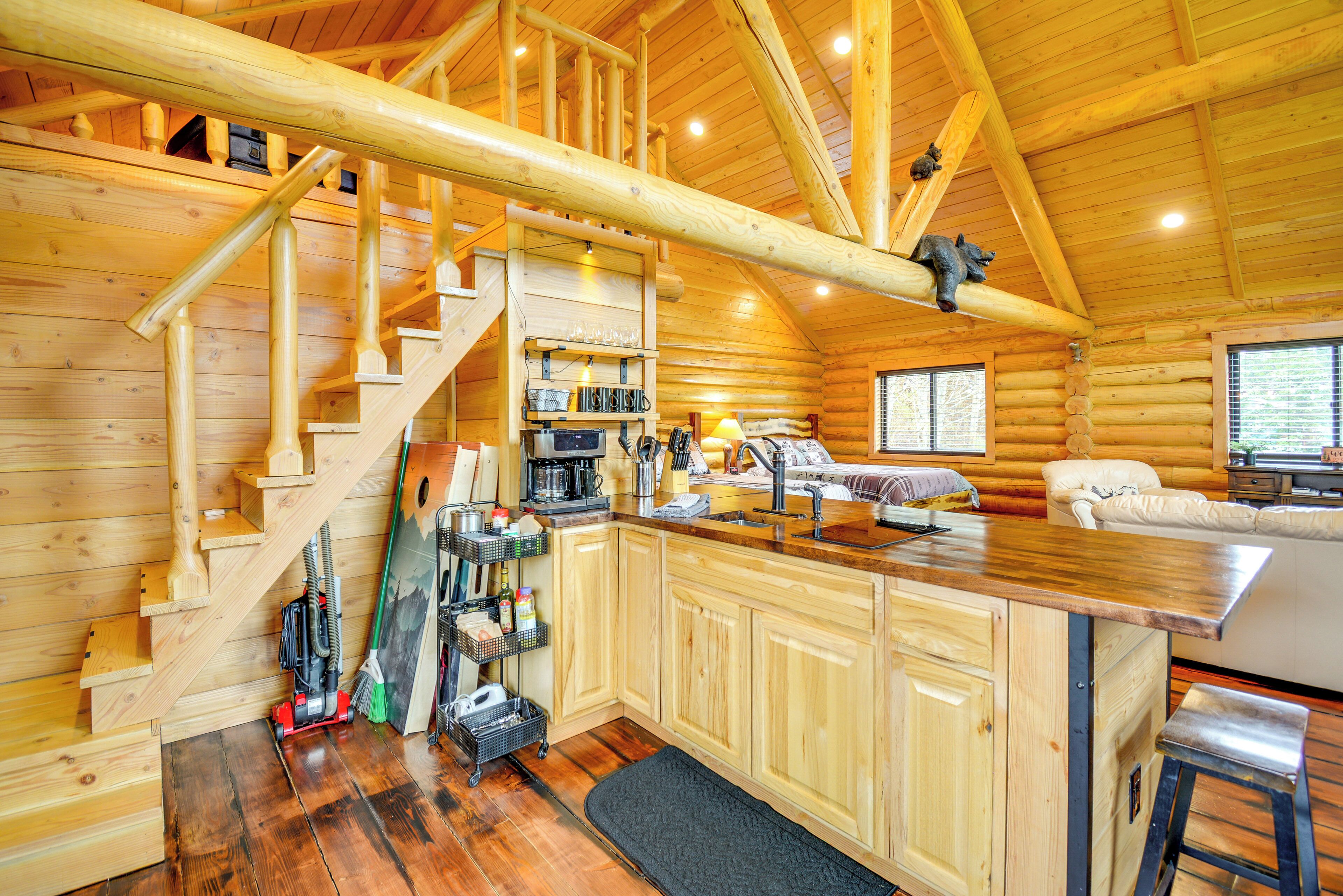 A log cabin interior with a wooden staircase, kitchen area, and a cozy seating area.