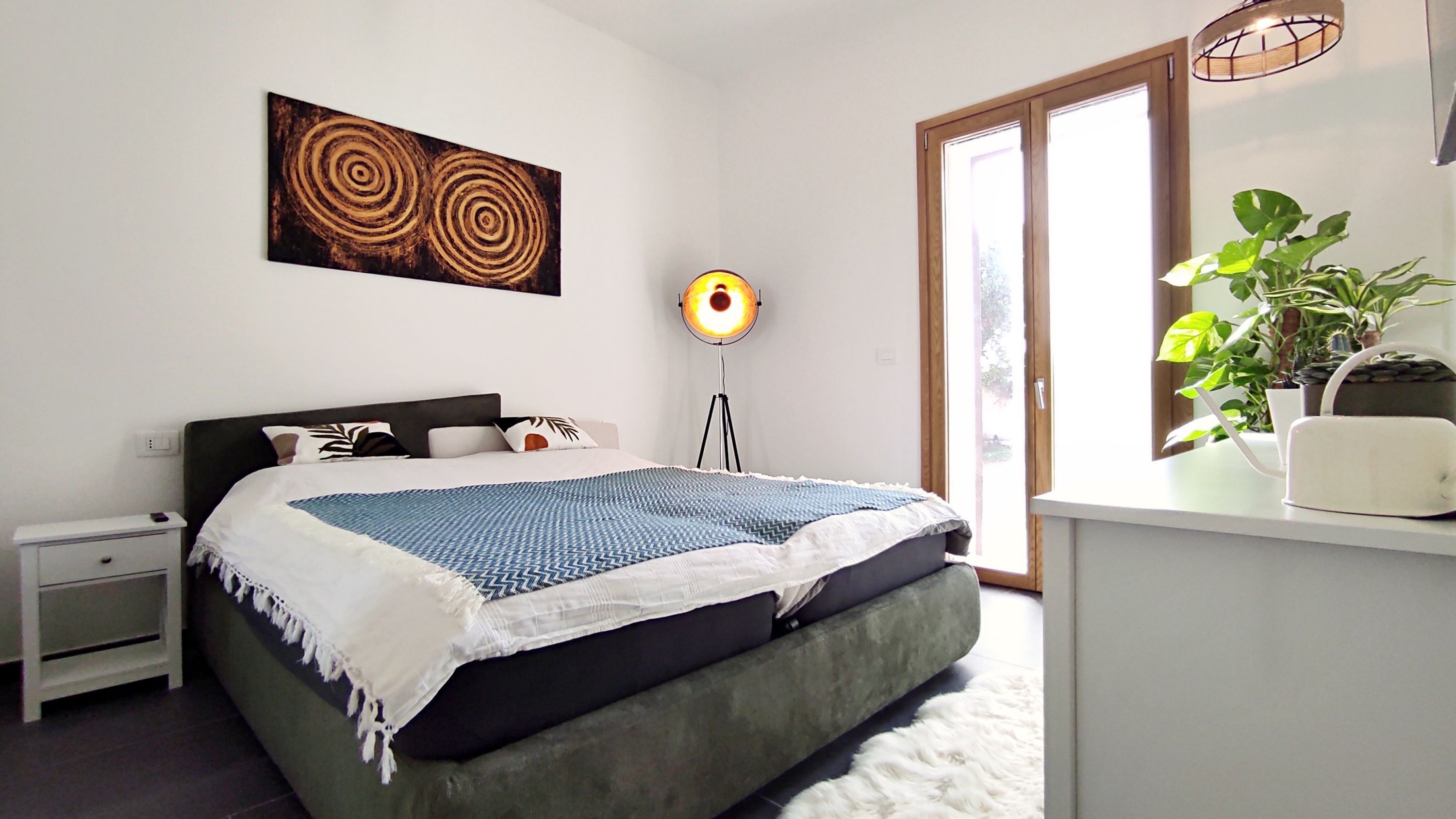 A bedroom with a bed, bedside table, floor lamp, and a window with a view.