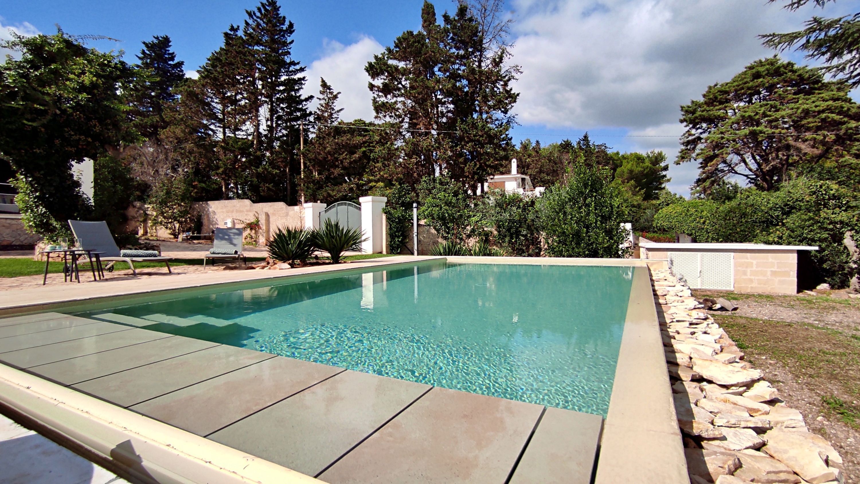 A rectangular swimming pool with a tiled deck, surrounded by a stone wall and various trees.