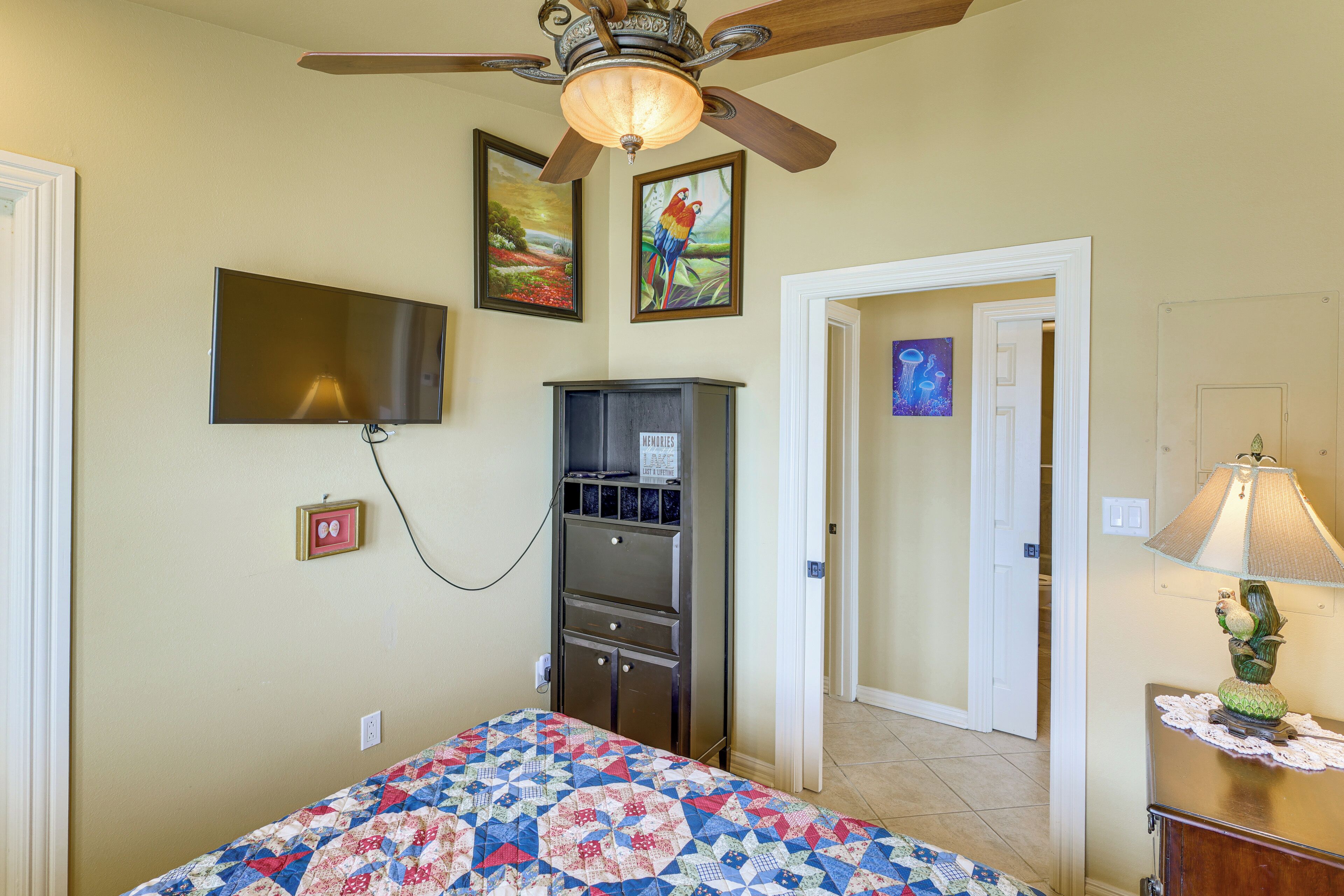 A bedroom with a bed, a television, a ceiling fan, a dresser, and a door leading to another room.