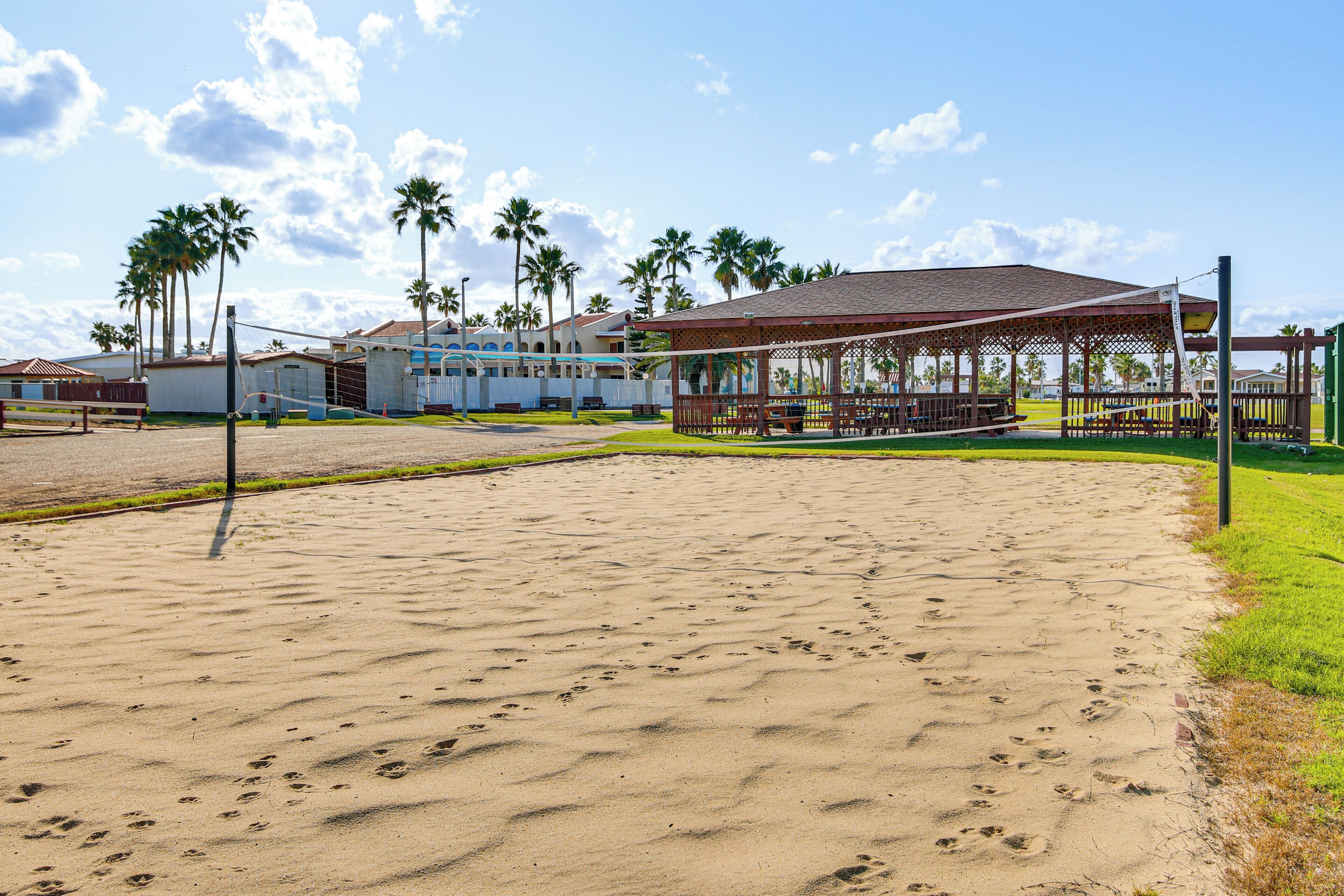 A sandy beach area with a pavilion, palm trees, and a building with a sloped roof.