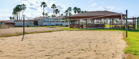 A sandy beach area with a pavilion, palm trees, and a building with a sloped roof.