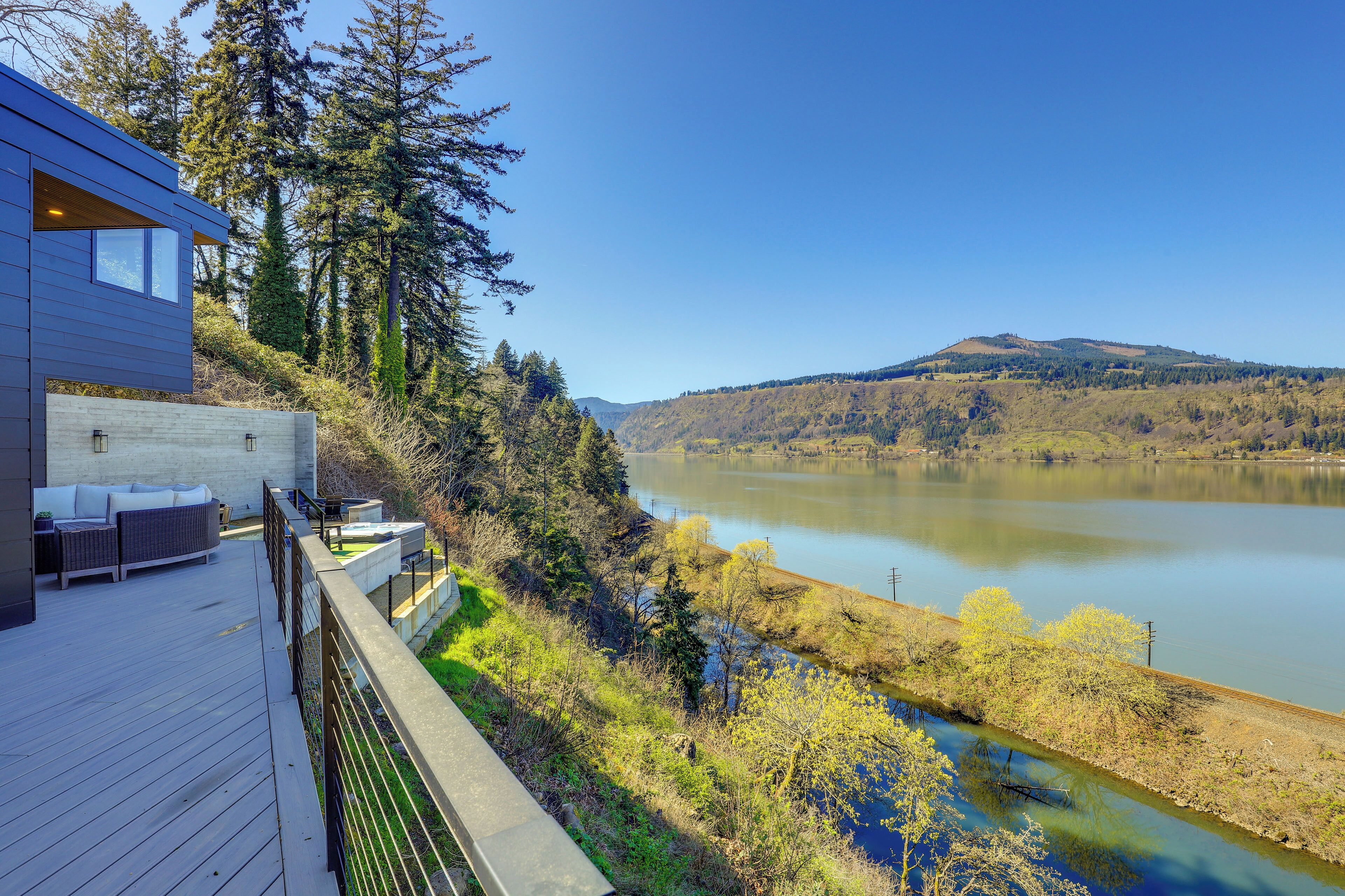 A modern house with a deck overlooking a river and forested hills.