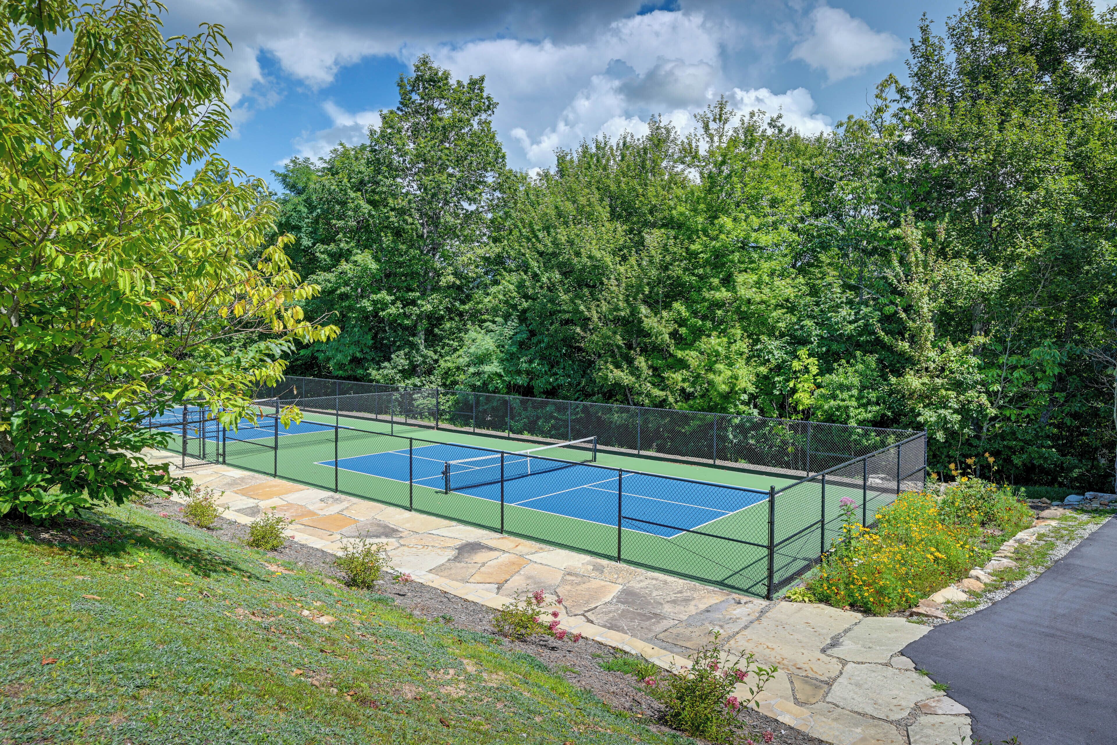 A tennis court surrounded by a stone pathway and a fence, with trees and flowers in the background.