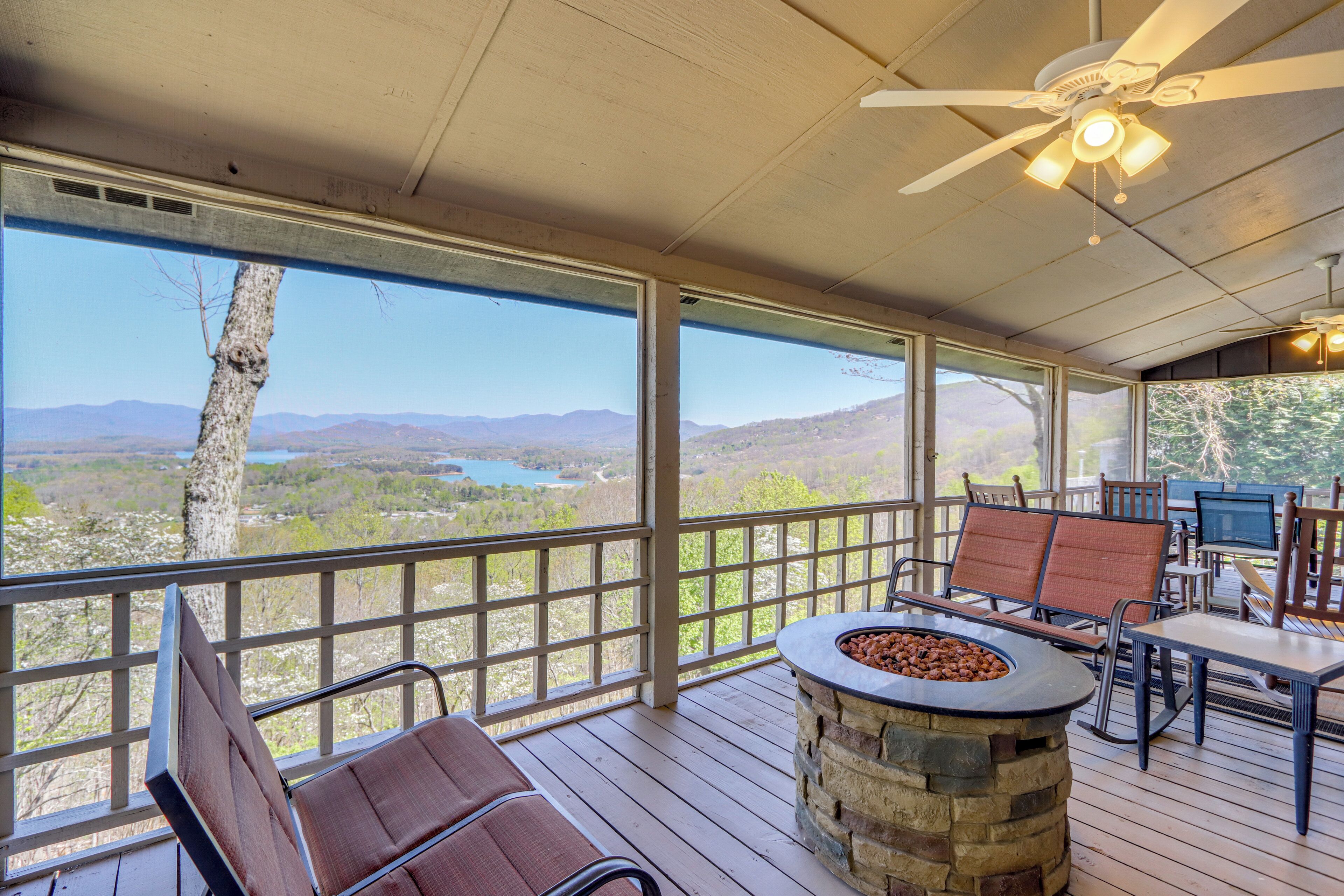 A covered outdoor deck with a stone fire pit, seating, and a view of a lake and mountains.