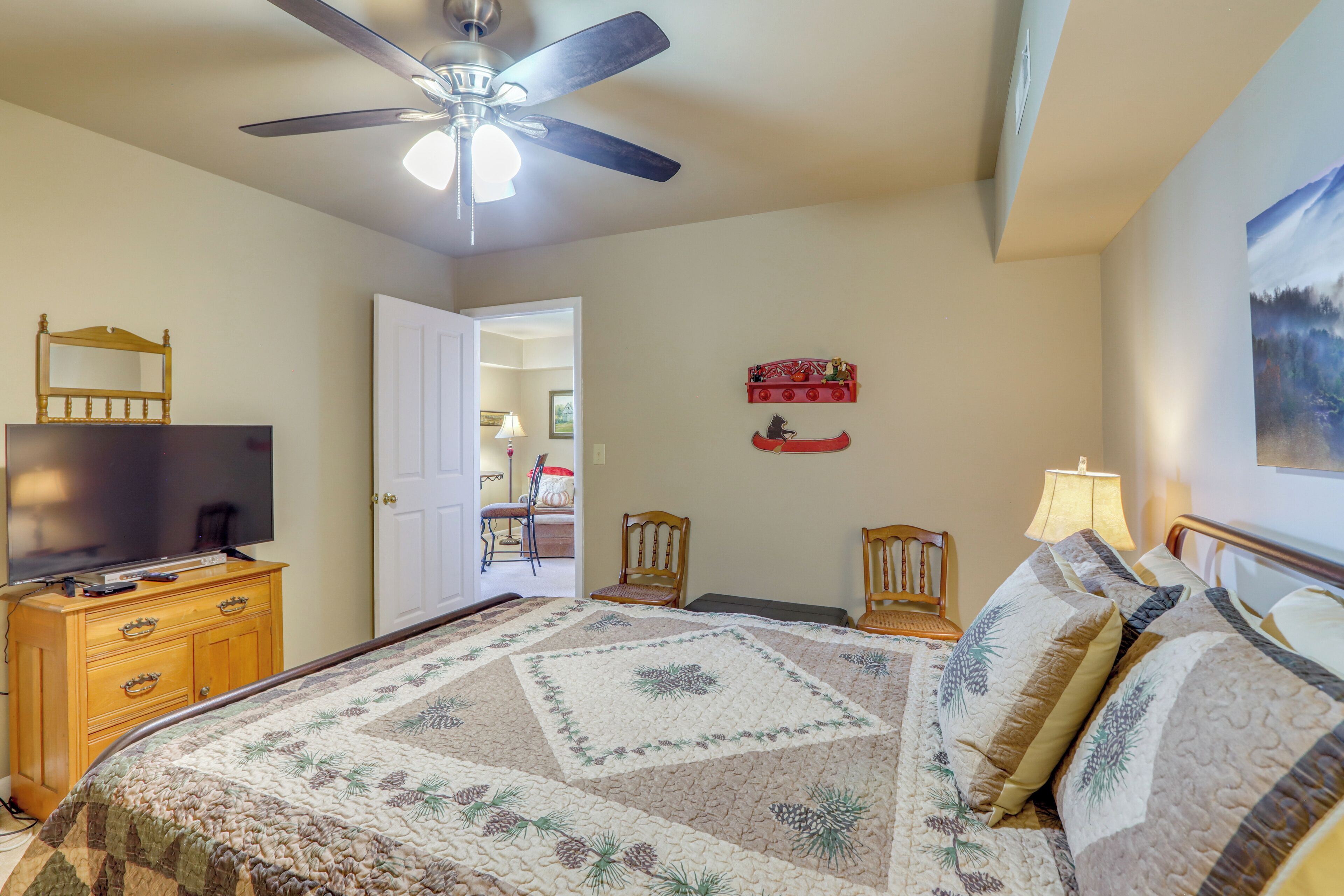 Une chambre à coucher avec un lit, un ventilateur de plafond, un téléviseur posé sur une commode en bois et une porte donnant sur une autre pièce.