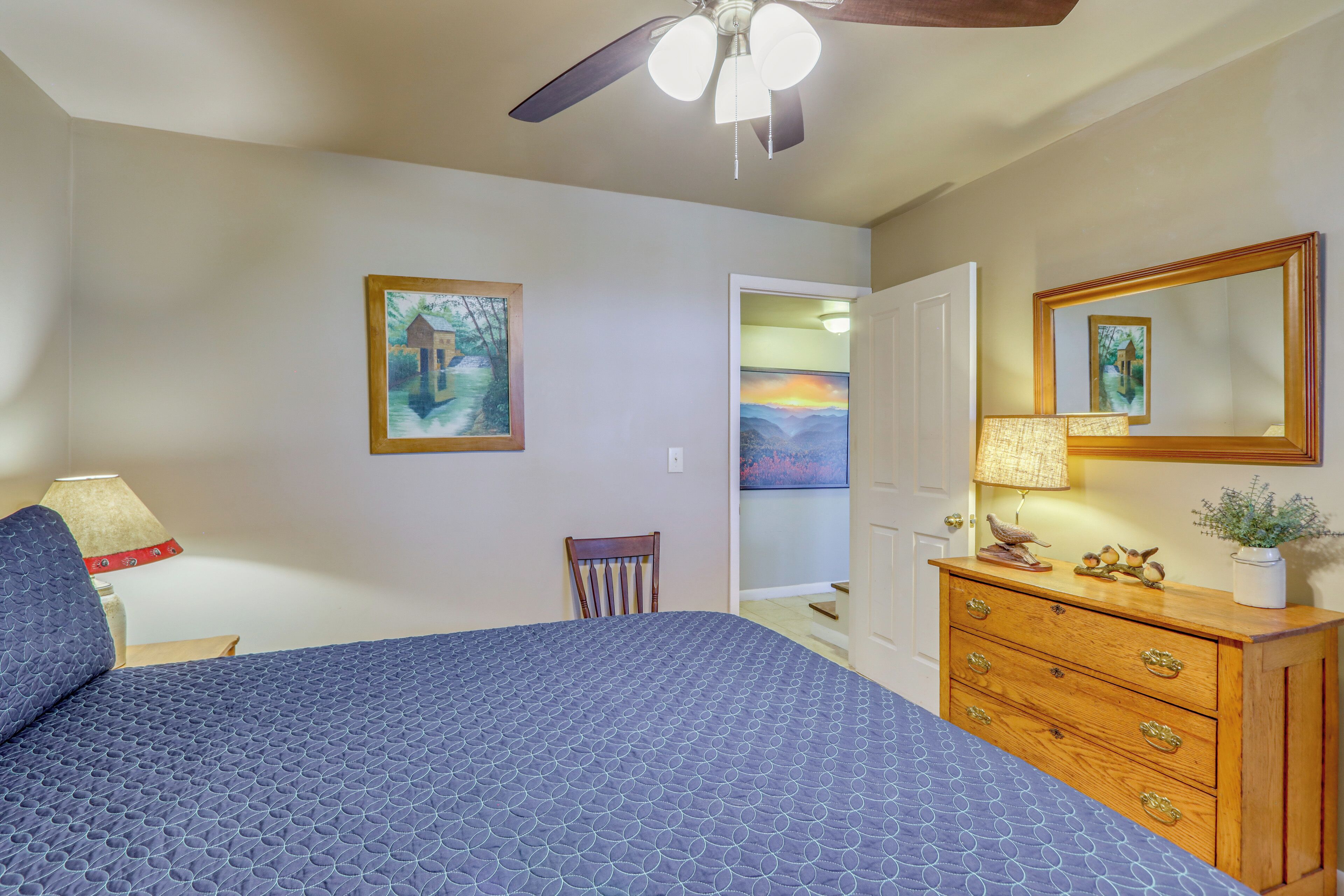 A bedroom with a bed, a dresser, a ceiling fan, a lamp, and two paintings on the wall.