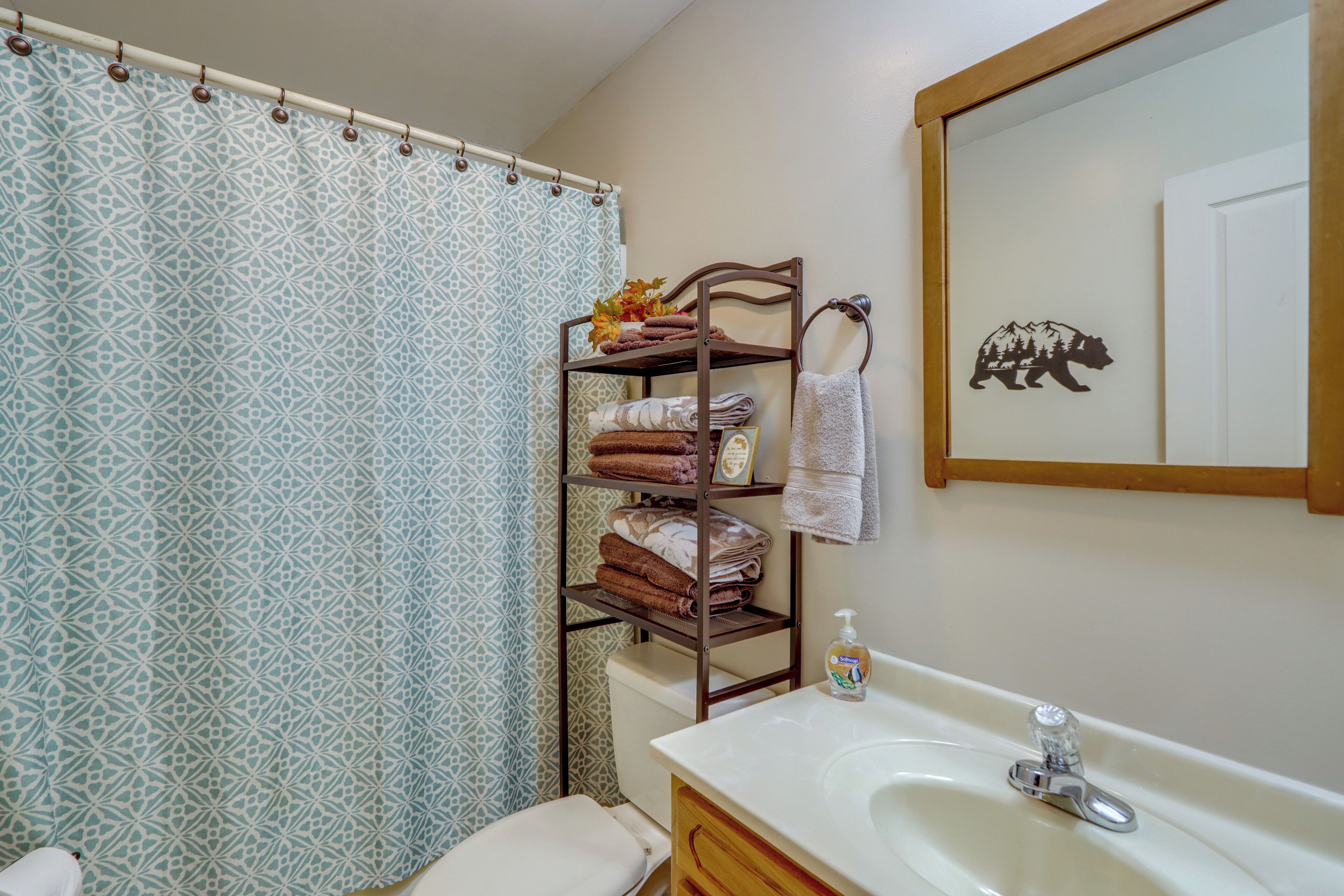 A bathroom with a patterned shower curtain, a wooden towel rack with folded towels, a sink with a soap dispenser, and a mirror with a bear silhouette.