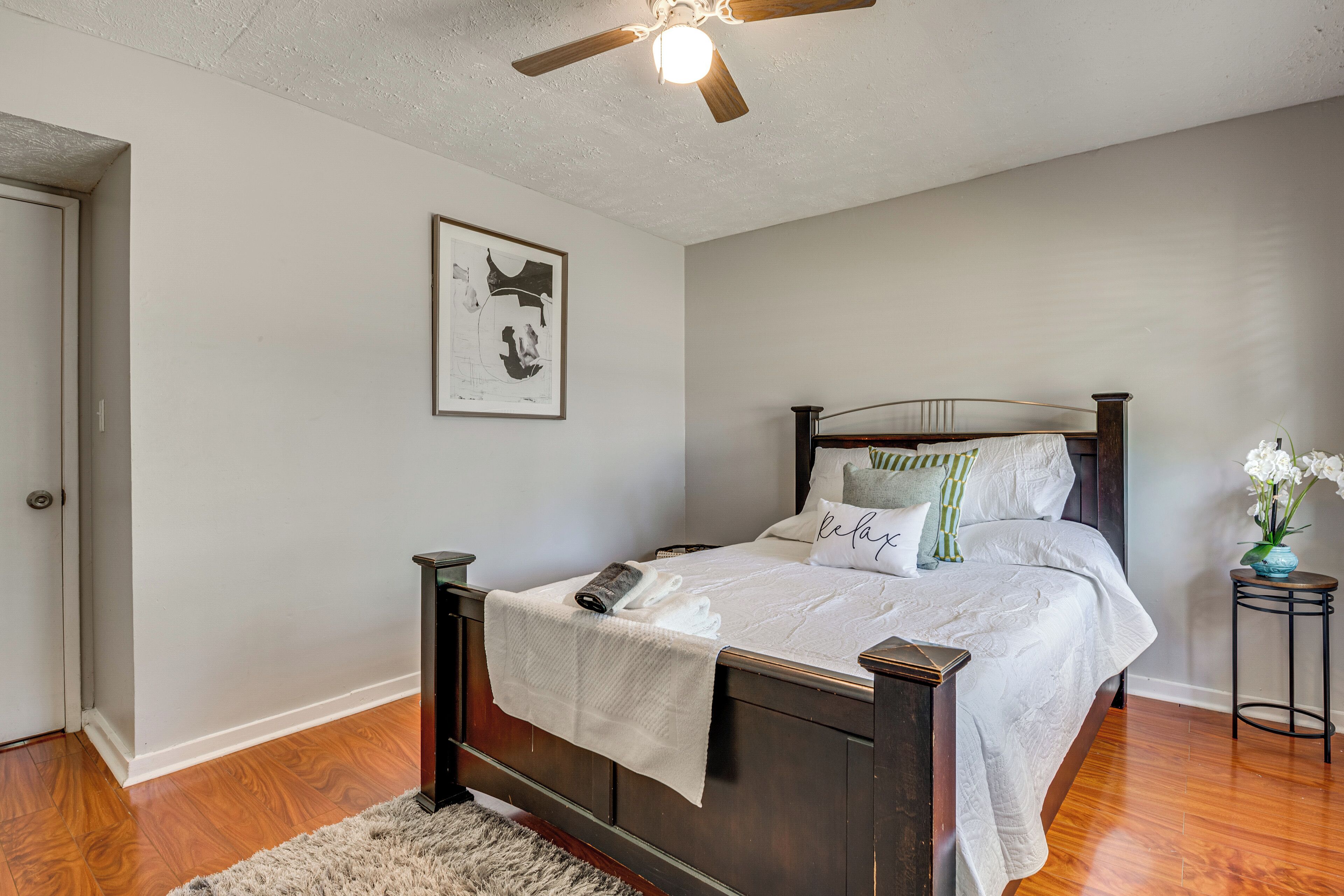 A bedroom with a wooden bed, a ceiling fan, a framed artwork, and a small side table with flowers.