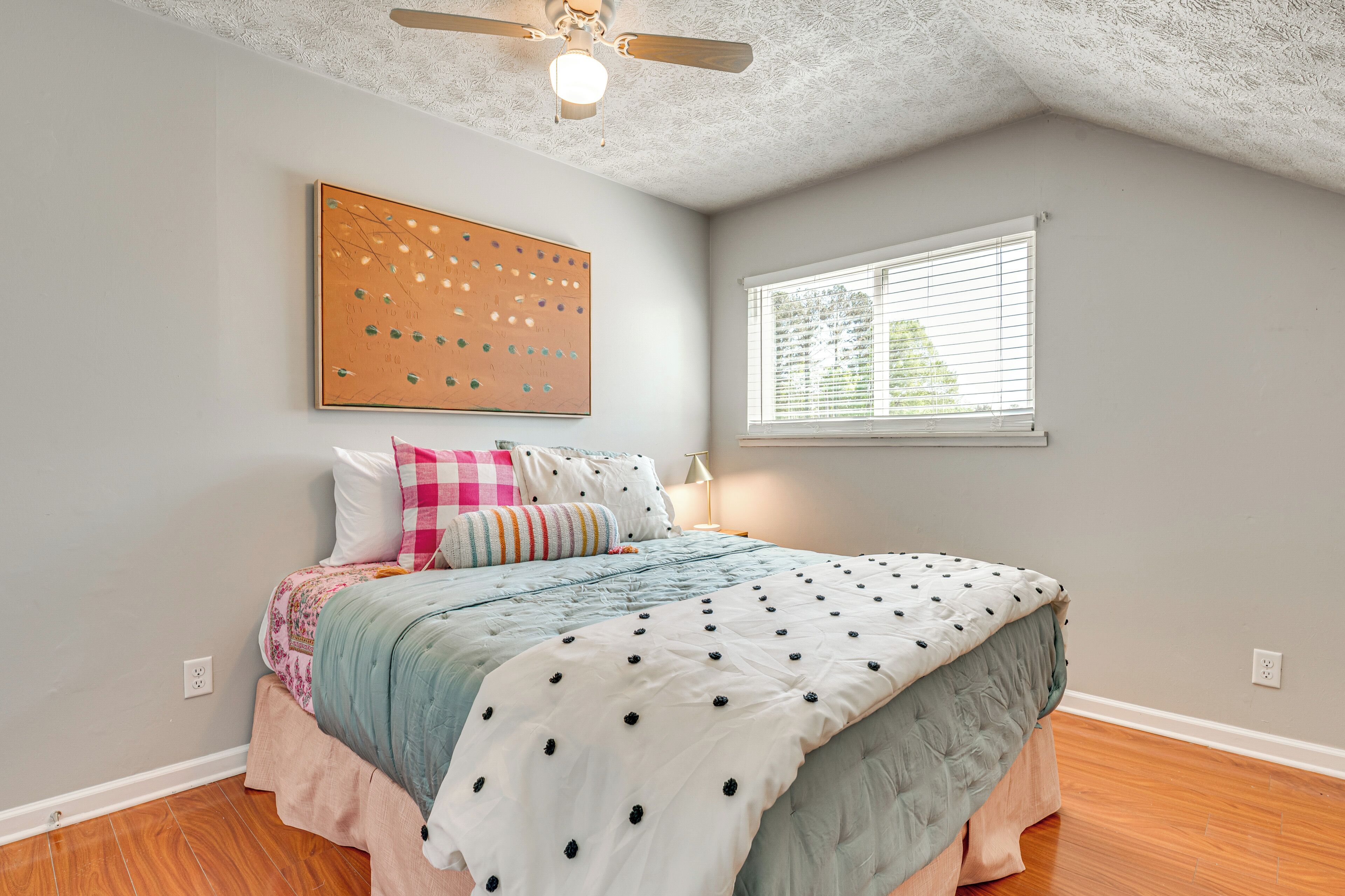 Un dormitorio con cama, ventilador de techo, ventana y una obra de arte en la pared.