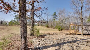 A rustic cabin with a wooden deck, surrounded by a natural landscape with trees and a dirt path. - Small Family Getaway in Mena w/ Trails On-site! (Mena)
