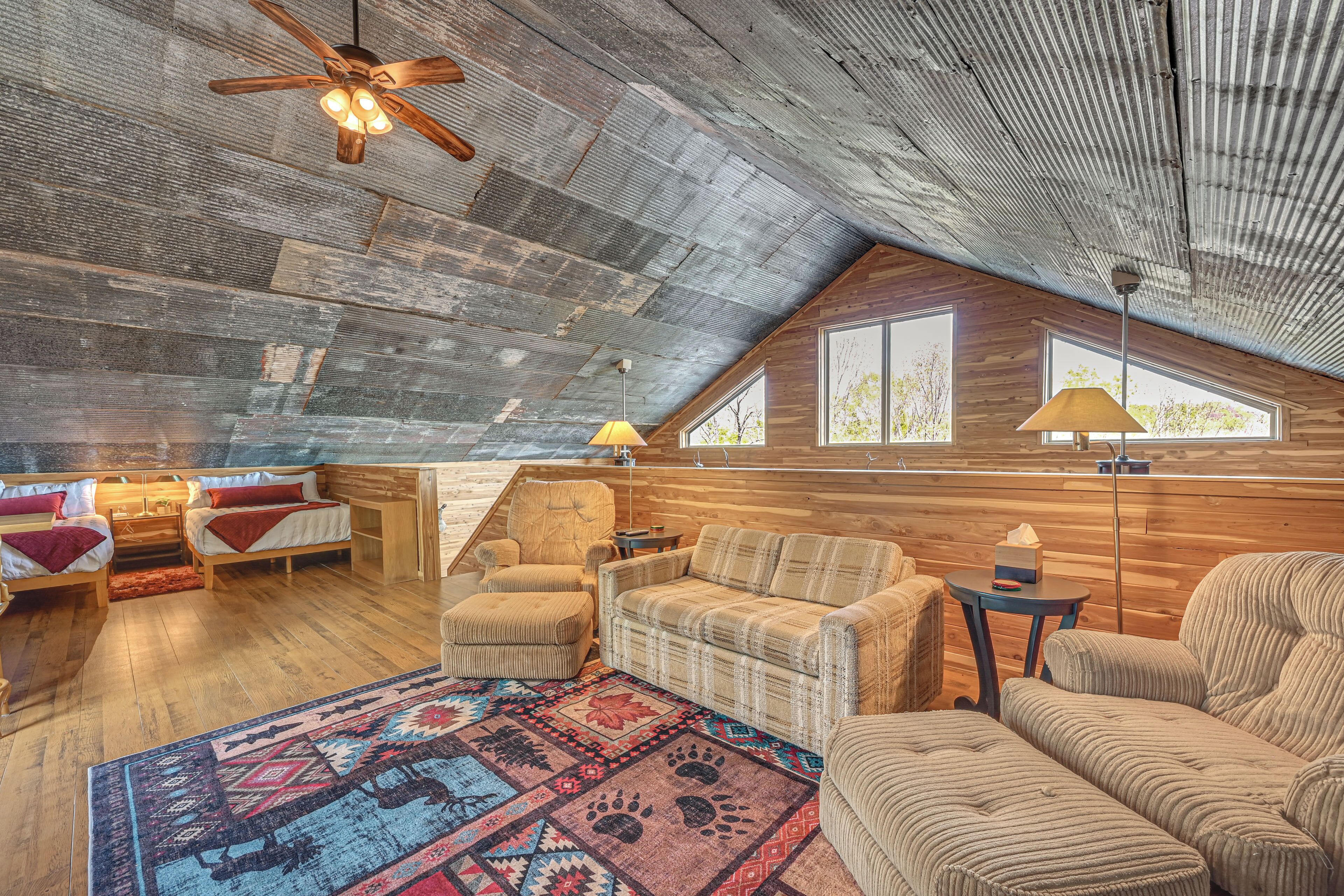 A cozy loft-style room with a sloped ceiling, wooden walls, a large window, and a variety of seating options including a sofa, chairs, and a bed.