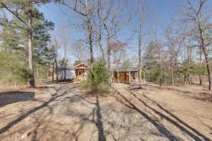 Une cabane rustique, accessible par un chemin de terre, entourée d’arbres et d’une clôture en bois.