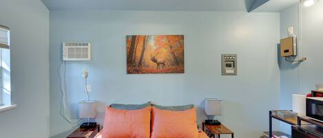 A bedroom with a bed, a painting, an air conditioning unit, a lamp, a desk, and a TV.