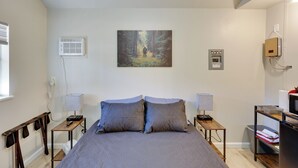 A bedroom with a bed, two bedside tables with lamps, a wall-mounted air conditioner, a painting, and a small cabinet. - Ouachitas Adventure Base! Charming Tiny Cabin (Mena)