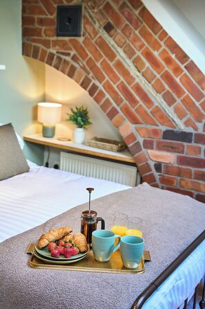 Un lit bien fait, avec un plateau de petit-déjeuner, un mur en briques et une petite table sur laquelle se trouvent une lampe et une plante.
