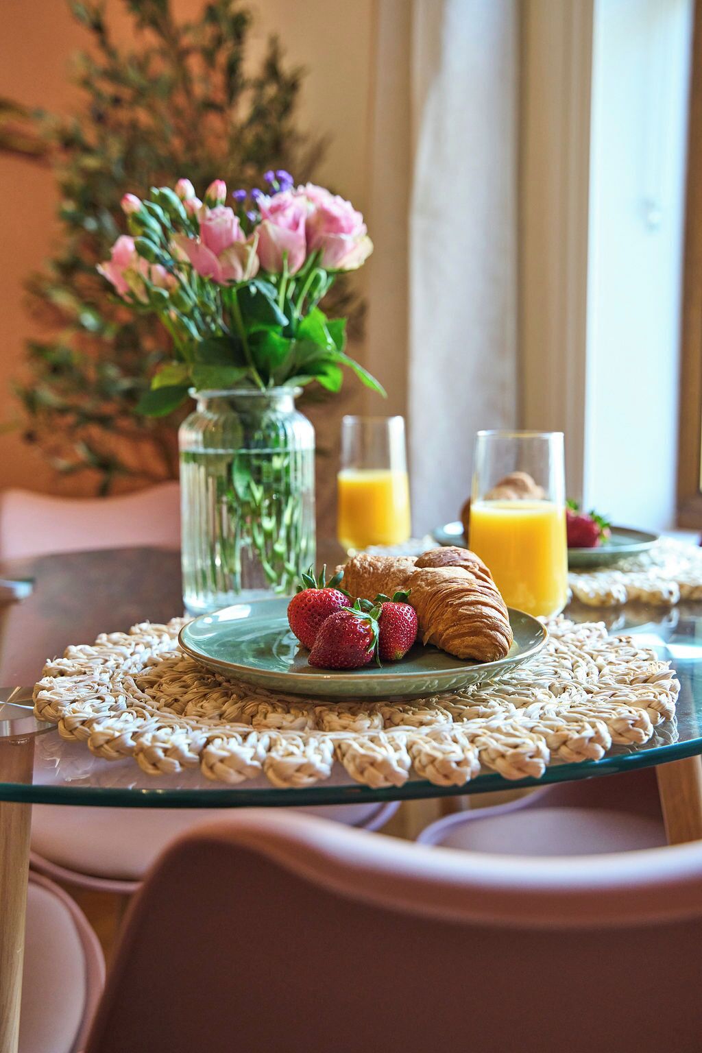 Une table dressée pour le petit-déjeuner, avec des croissants, des fraises et du jus d’orange, le tout posé sur une table en verre surmontée d’un set de table en fibres tressées.