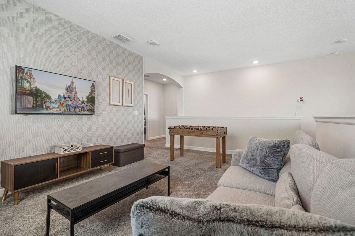 A living room with a sofa, a coffee table, a wooden cabinet, a foosball table, and a wall-mounted TV.