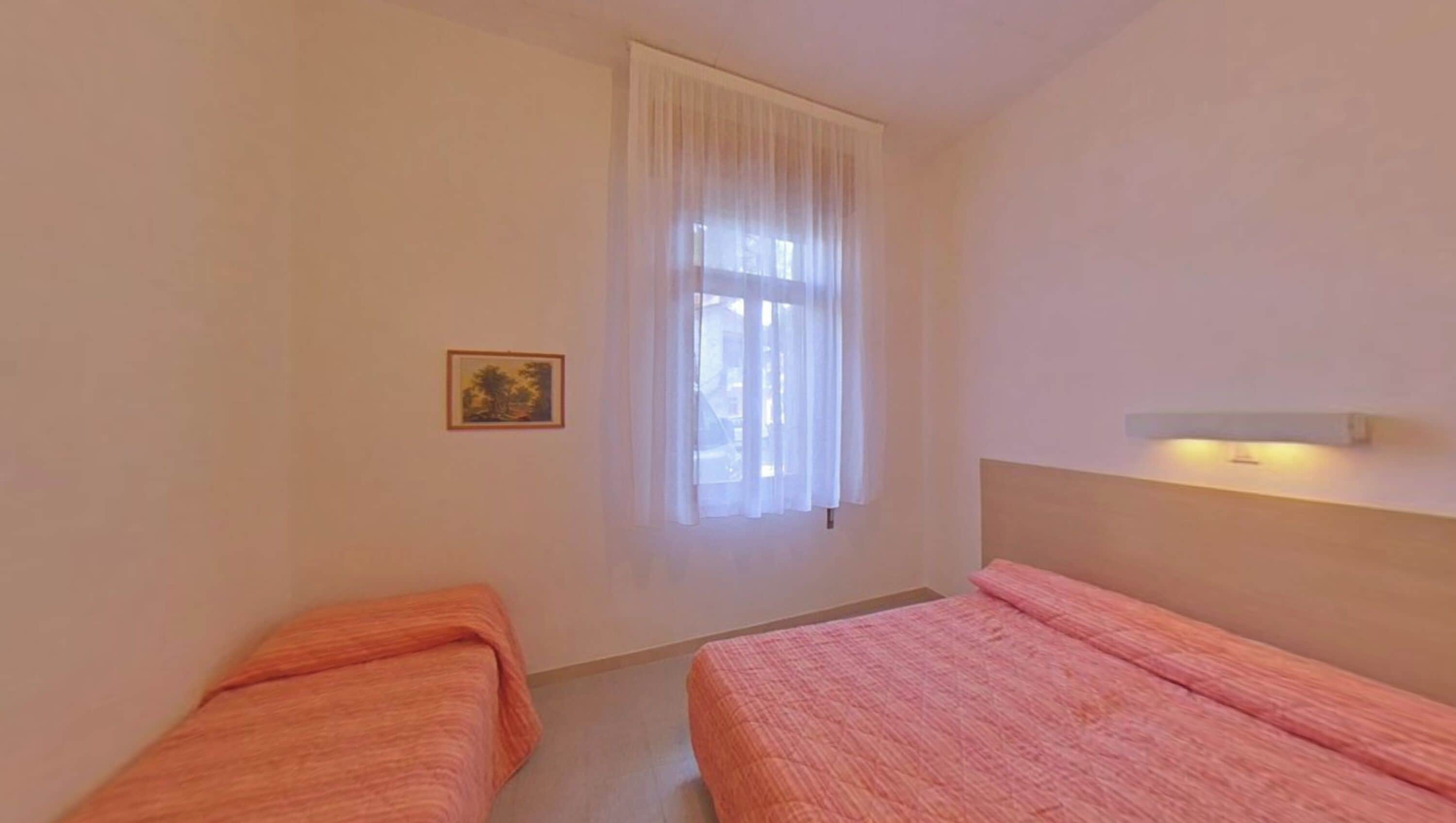 Ein Zimmer mit zwei Betten, einem Fenster mit transparenten Vorhängen, einem gerahmten Bild an der Wand und einem Nachttisch mit Lampe.