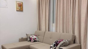 Ein Wohnzimmer mit Ecksofa, einem kleinen weißen Tisch, einem gemusterten Teppich und einem Fenster mit Vorhängen.