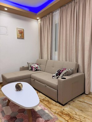 A living room with a sectional sofa, a small white table, a patterned rug, and a window with curtains. - Charming 2-bedroom Apartment in the Heart of Hurghada (Hurghada)