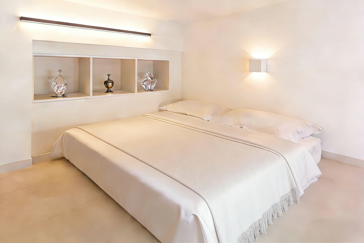 A bedroom with a large bed, a bedside table, a wall-mounted shelf with decorative items, and a wall-mounted light.