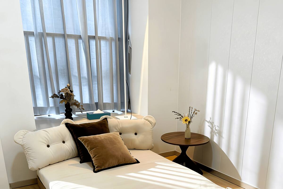 A modern bedroom with a bed, a bedside table, a window with curtains, and a vase with flowers.