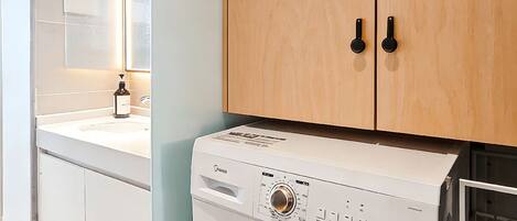 A modern laundry room with a front-loading washing machine, wooden cabinets, and a mirror.