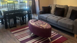 Een woonkamer met een bank, voetenbankje, gestreept tapijt, een eettafel met stoelen en een raam met gordijnen.