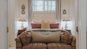 A bedroom with a bed, two bedside lamps, a ceiling fan, a window with curtains, and a plaid sofa. - Charming cottage on 8 acres of peace & tranquility just a short drive from town. (Boaz)