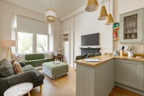 A modern living room with a green sofa, a small white table, and a kitchen area with a wooden countertop and a television. - Stockbridge ‘Little Gem’ Flat, Sleeps 3 (Stockbridge)