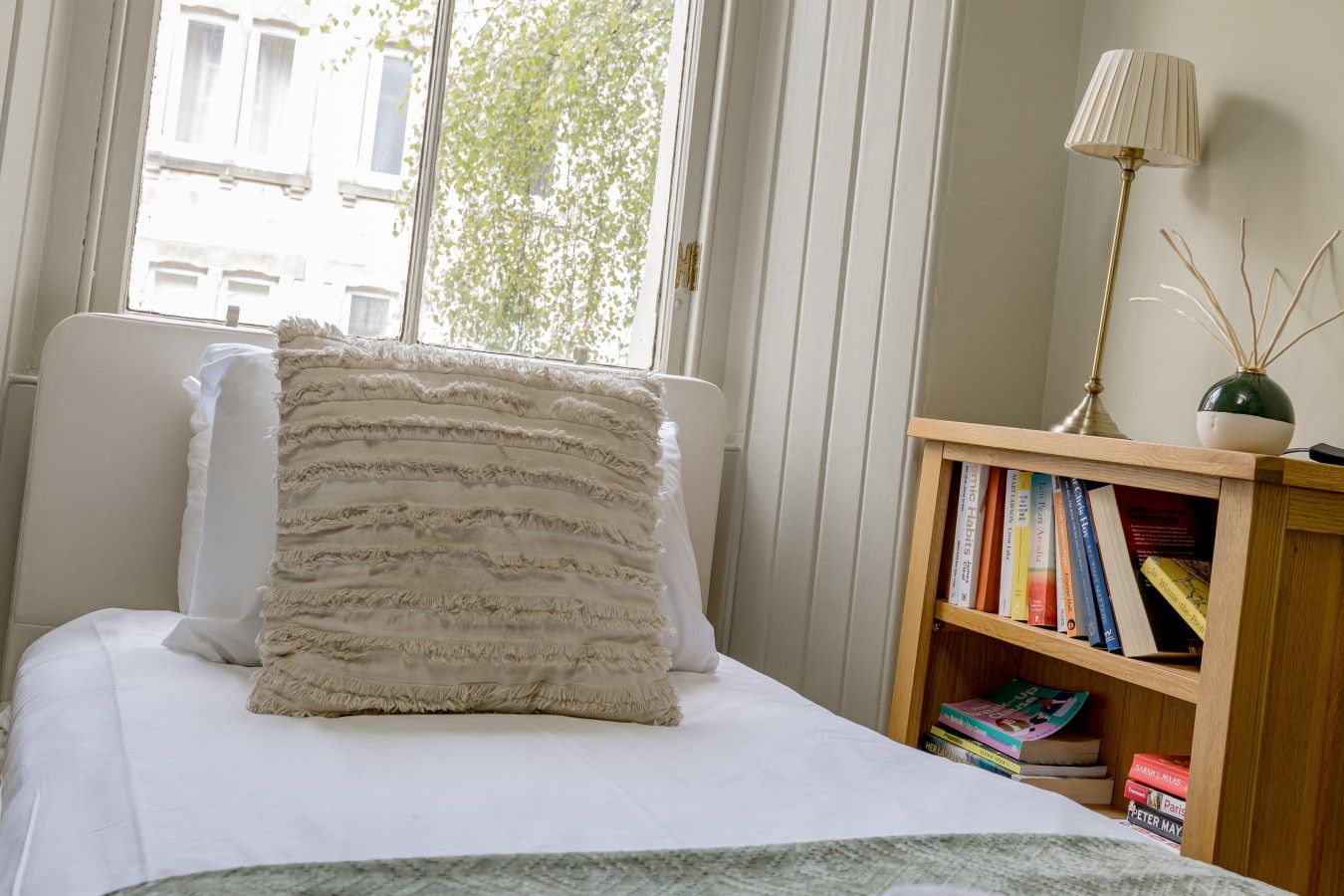 A bedroom with a bed, a bedside lamp, a bookshelf, and a window with a view of buildings.