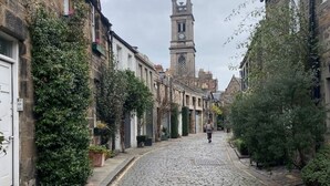 Une rue pavée bordée de bâtiments historiques en pierre, un clocher visible au loin et une personne qui marche dans la rue.