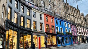 Une rue pavée bordée de bâtiments aux couleurs vives, une boutique bien visible appelée « LADESWALAR », et un bâtiment historique doté d’une tour horlogée.