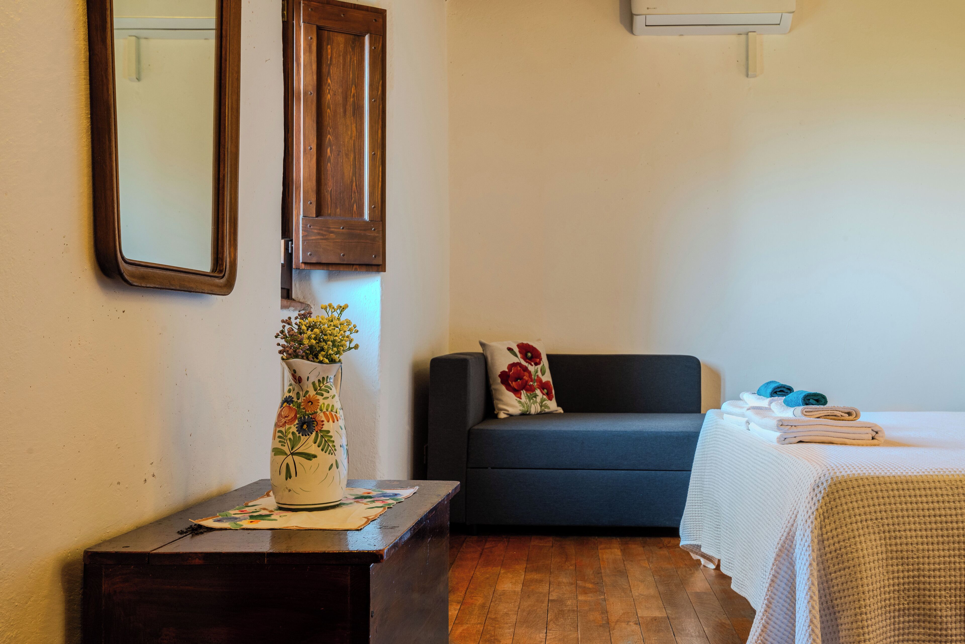 A room with a wooden dresser, a dark sofa, a bed with a white bedspread, a vase with flowers, and a mirror on the wall.