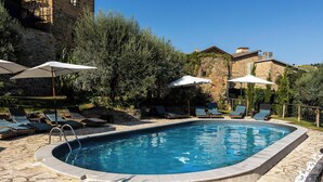 Una piscina con sillones de descanso y sombrillas, rodeada de muros de piedra y vegetación.