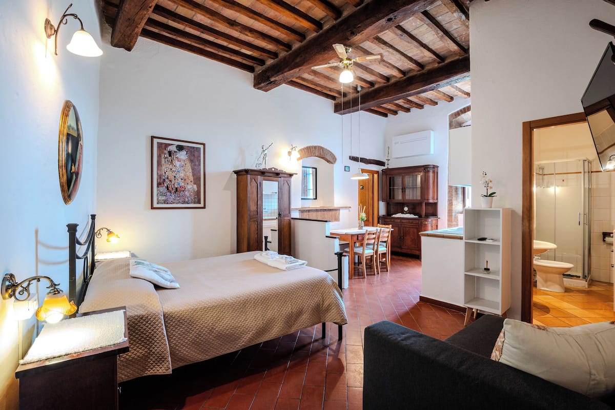 A bedroom with a bed, bedside table, lamp, and a view into the kitchen.