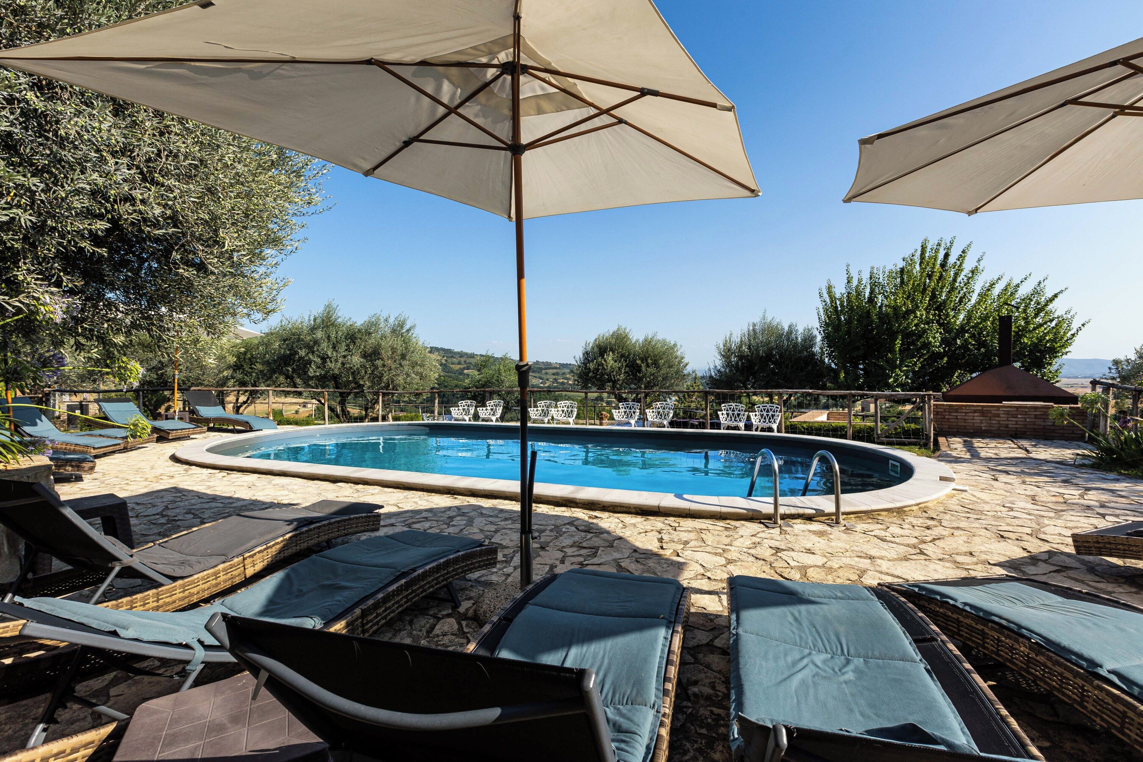 Piscina com espreguiçadeiras e guarda-sóis, cercada por chão de pedra e vegetação.