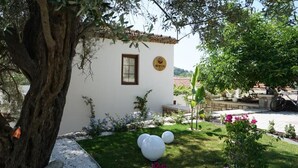 A white building with a window and a sign, surrounded by a well-maintained garden with grass, white spherical objects, and flowering plants. - Muyo House (Urla)