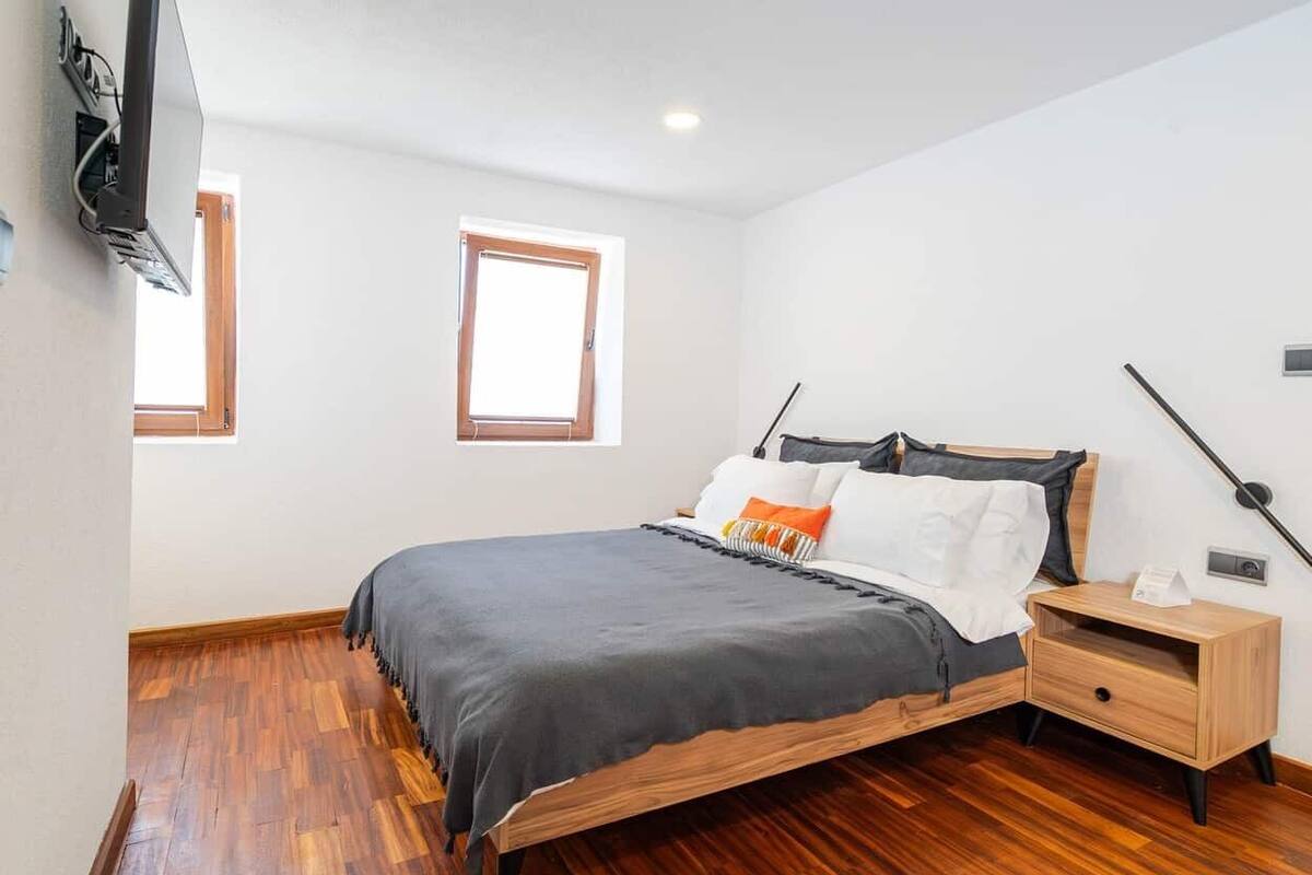 Quarto com cama de madeira, mesa de cabeceira, televisão na parede e janela.
