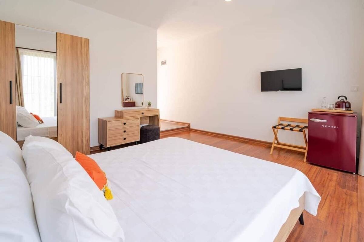 Quarto de hotel com uma cama grande, um guarda-roupa de madeira, uma pequena escrivaninha com espelho, televisão na parede e um minibar vermelho.