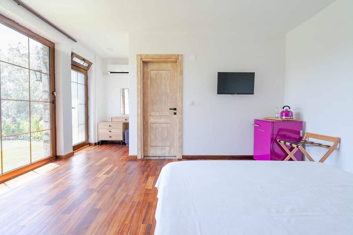 Um quarto com uma cama, uma porta de madeira, uma televisão, um armário rosa e uma janela grande.
