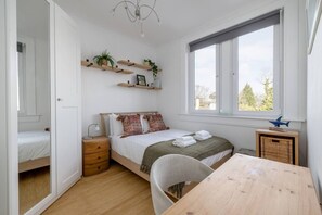 Ett sovrum med en säng, ett nattduksbord, en lampa, ett trägolv, ett fönster och utsikt över träd.