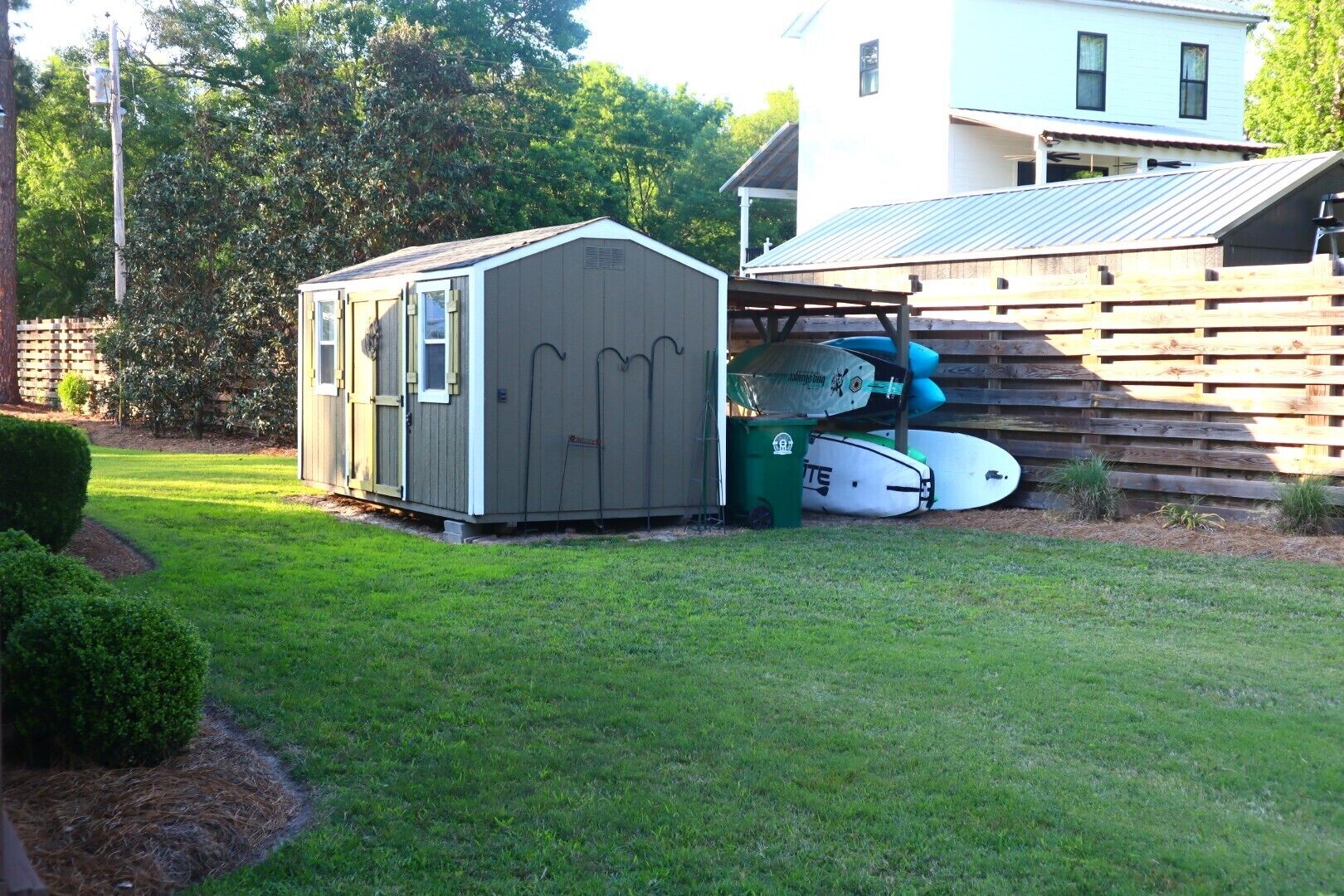 A small shed with a door and windows, a fenced area with surfboards, and a house in the background.