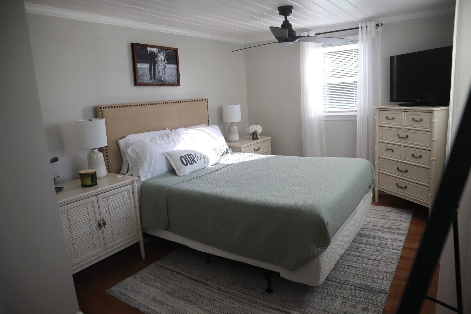 A bedroom with a bed, nightstands, a television, a ceiling fan, and a window with blinds.