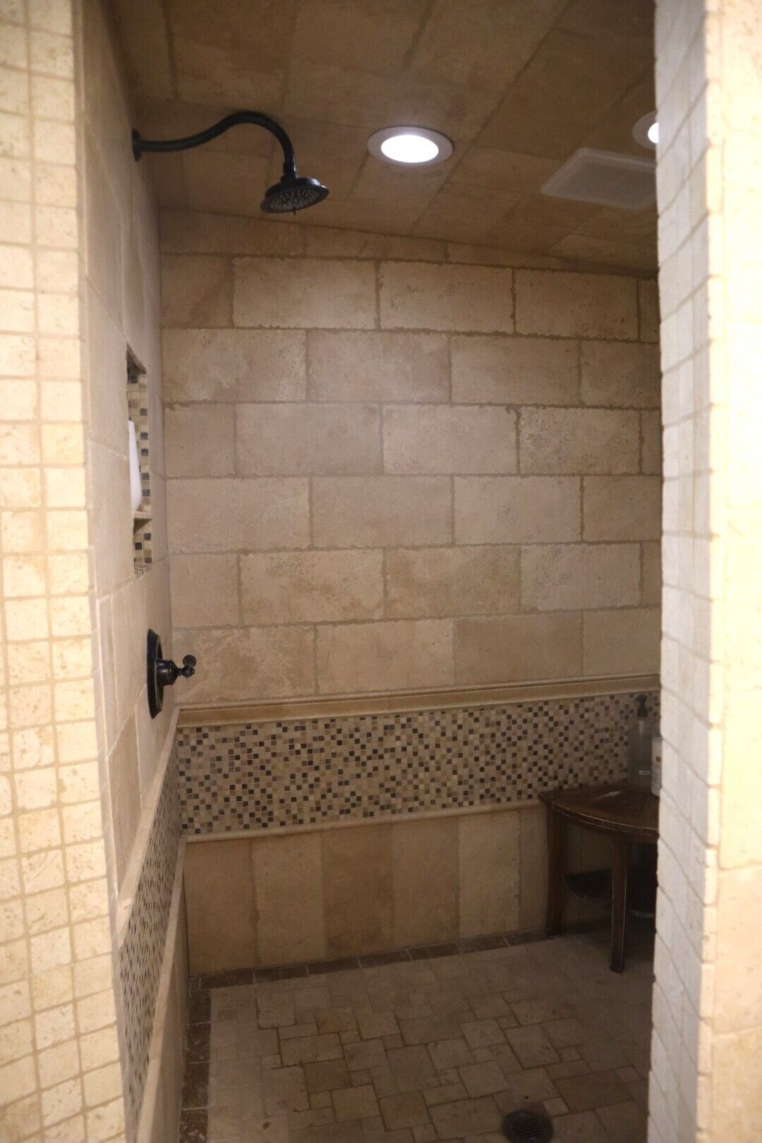 A narrow shower area with tiled walls, a tiled floor, a showerhead, and a small wooden bench.